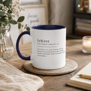 May include: A white ceramic mug with a dark blue interior and handle. The mug features black printed text that reads: "Lehrer [Herr Wagner] Substantiv, m. Ein Lehrer ist eine Person, die aus kleinen Fortschritten gro&szlig;e Erfolge macht und dabei jeden einzelnen Schritt wertsch&auml;tzt."