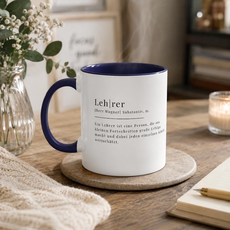 May include: A white ceramic mug with a dark blue interior and handle. The mug features black printed text that reads: "Lehrer [Herr Wagner] Substantiv, m. Ein Lehrer ist eine Person, die aus kleinen Fortschritten gro&szlig;e Erfolge macht und dabei jeden einzelnen Schritt wertsch&auml;tzt."
