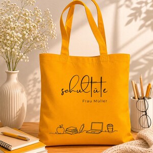 K&ouml;nnte beinhalten: Senfgelbe Tragetasche mit schwarzer Schrift, die "Schult&uuml;te" und "Frau M&uuml;ller" lautet. Die Tasche zeigt eine Strichzeichnung eines Apfels, B&uuml;cher, eines Stifts, eines Laptops und einer Kaffeetasse. Die Tasche hat zwei Henkel.