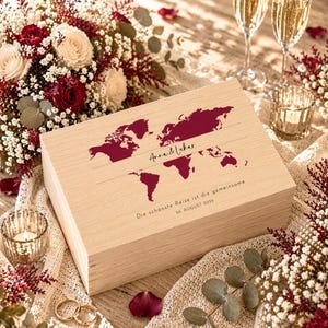 May include: A light-colored wooden box with a world map design in burgundy. The box is inscribed with the names "Anna & Lukas" and the date "26. AUGUST 2026". The box is surrounded by flowers, candles, and champagne glasses.