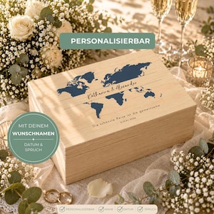 May include: A light-colored wooden box with a blue world map design and the names "Katharina & Alexander." The text "Die sch&ouml;nste Reise ist die gemeinsame" is also visible. The box is surrounded by flowers and wedding rings.