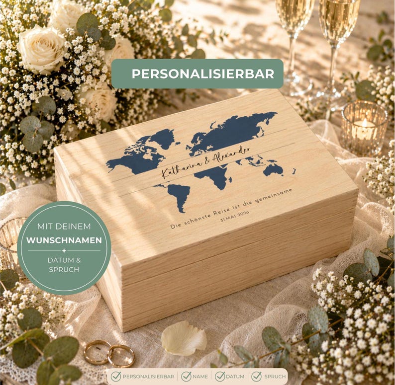 May include: A light-colored wooden box with a blue world map design and the names "Katharina & Alexander." The text "Die sch&ouml;nste Reise ist die gemeinsame" is also visible. The box is surrounded by flowers and wedding rings.