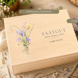 Könnte beinhalten: Eine rechteckige, helle Holzbox mit einer Aquarellillustration von Wildblumen auf der linken Seite. Der Text lautet SAATGUT, Blumen-Kräuter-Gemüse und Familie MÜLLER. Zwei rechteckige Etiketten über der Box zeigen PERSONALISIERBAR und 6 MOTIVE.