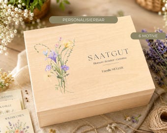 Può includere: Una scatola rettangolare in legno chiaro con un'illustrazione ad acquerello di fiori di campo sul lato sinistro. Il testo riporta SAATGUT, Blumen-Kräuter-Gemüse e Familie MÜLLER. Due etichette rettangolari sopra la scatola indicano PERSONALISIERBAR e 6 MOTIVE.