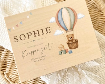 Könnte beinhalten: Eine rechteckige Holzbox mit einem Aufdruck eines Teddybären im Heißluftballon, bunten Bauklötzen und Wimpelketten. Der Text lautet SOPHIE, Erinnerungen an meine Krippenzeit, Mäusegruppe und 2024-2026.