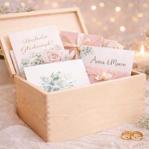 May include: A light wooden box filled with wedding stationery. The box contains cards with floral designs and text, along with pink envelopes tied with ribbons. Gold wedding rings are in the foreground.