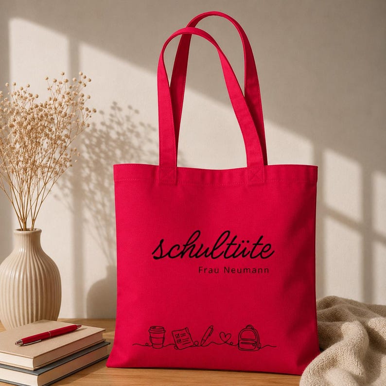 K&ouml;nnte beinhalten: Eine leuchtend rote Tragetasche mit schwarzem Text, der "schult&uuml;te Frau Neumann" lautet. Die Tasche zeigt eine Strichzeichnung einer Kaffeetasse, eines Notizblocks, eines Bleistifts, eines Herzens und eines Rucksacks. Die Tasche hat zwei Griffe.