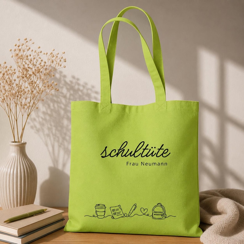 K&ouml;nnte beinhalten: Eine hellgr&uuml;ne Tragetasche mit schwarzem Text "schult&uuml;te Frau Neumann". Darunter eine Strichzeichnung einer Kaffeetasse, eines Notizblocks, eines Stifts, eines Herzens und eines Rucksacks. Die Tasche hat zwei Henkel.