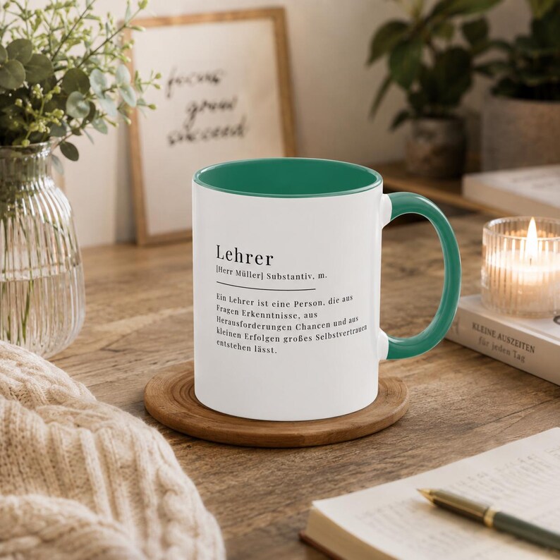 May include: A white ceramic mug with a dark green interior and handle, featuring black printed text that defines the German word Lehrer, describing a person who fosters self-confidence through questions, challenges, and small successes.