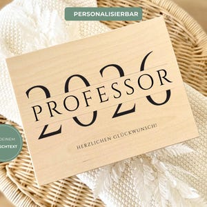 Könnte beinhalten: Eine hellfarbene Holztafel mit dem Wort "PROFESSOR" in großen schwarzen Buchstaben, mit den Zahlen "2026" darüber und darunter. Der Text "HERZLICHEN GLÜCKWUNSCH!" steht darunter. Die Tafel liegt auf einer gewebten Oberfläche.