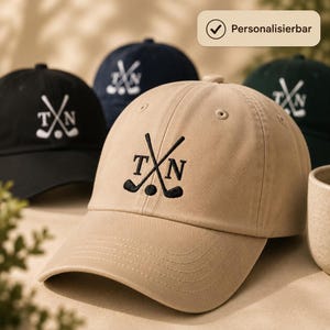 Könnte beinhalten: Eine beigefarbene Baseballkappe mit einer Stickerei aus zwei gekreuzten Golfschlägern, einem Golfball und den Buchstaben T und N. Die Kappe hat einen gebogenen Schirm und gestickte Ösen. Weitere Baseballkappen in Schwarz, Marineblau und Dunkelgrün sind zu sehen.