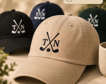 Può includere: Un cappellino da baseball color beige con un ricamo raffigurante due mazze da golf incrociate, una pallina da golf e le lettere T e N. Il cappellino presenta una visiera curva e occhielli cuciti. Sono visibili altri cappellini neri, blu navy e verde scuro.