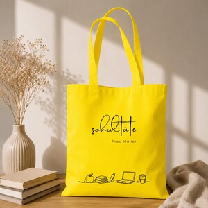 K&ouml;nnte beinhalten: Eine leuchtend gelbe Tragetasche mit schwarzer Schrift und einer Grafik von Apfel, B&uuml;chern, Stift, Laptop und Kaffeetasse. Die Tasche hat zwei Henkel und den Text "Schult&uuml;te Frau M&uuml;ller".