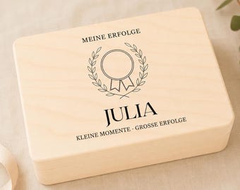 Può includere: Una scatola rettangolare in legno chiaro con coperchio incernierato. Il coperchio presenta una scritta nera che recita MEINE ERFOLGE, il nome JULIA e KLEINE MOMENTE · GROSSE ERFOLGE, insieme a un grafico di una medaglia circolare con foglie di alloro.