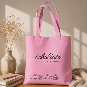 K&ouml;nnte beinhalten: Eine rosa Tragetasche mit dem deutschen Wort "schult&uuml;te" in schwarzer Schrift. Darunter steht "Frau Neumann". Am unteren Rand befindet sich eine Strichzeichnung einer Kaffeetasse, eines Notizblocks, eines Stifts, eines Herzens und eines Rucksacks.