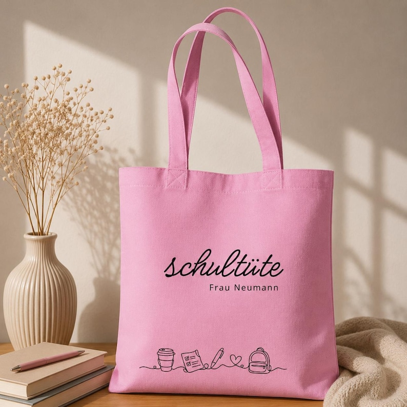 K&ouml;nnte beinhalten: Eine rosa Tragetasche mit dem deutschen Wort "schult&uuml;te" in schwarzer Schrift. Darunter steht "Frau Neumann". Am unteren Rand befindet sich eine Strichzeichnung einer Kaffeetasse, eines Notizblocks, eines Stifts, eines Herzens und eines Rucksacks.