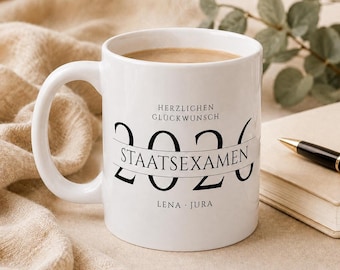 Può includere: Una tazza in ceramica bianca con testo nero che recita HERZLICHEN GLÜCKWUNSCH, 2026, STAATSEXAMEN e LENA JURA. La tazza ha un manico grande ed è riempita con un liquido marrone chiaro. Il design include un banner orizzontale sull'anno 2026.