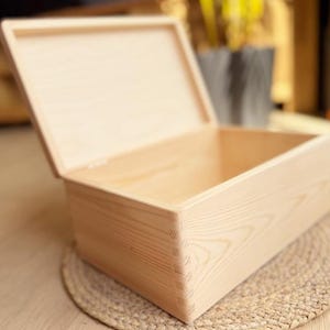 May include: A light-colored wooden box with a hinged lid, partially open. The box is rectangular with dovetail joints at the corners. The box is sitting on a woven mat.