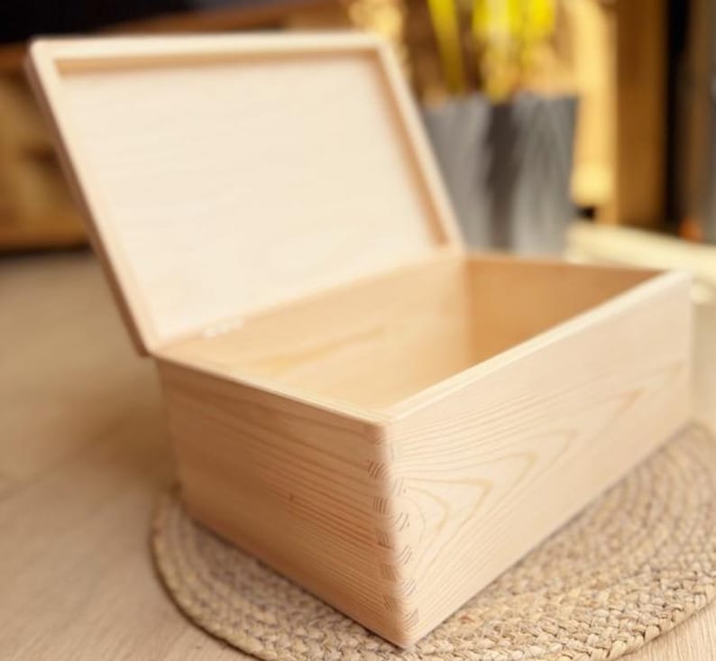 May include: A light-colored wooden box with a hinged lid, partially open. The box is rectangular with dovetail joints at the corners. The box is sitting on a woven mat.