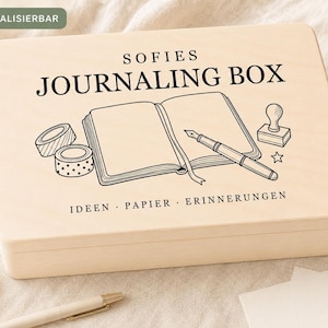 Könnte beinhalten: Eine rechteckige, helle Holzbox mit Klappdeckel und schwarzem Aufdruck mit dem Text SOFIES JOURNALING BOX sowie IDEEN PAPIER ERINNERUNGEN. Die Grafik auf dem Deckel zeigt ein offenes Notizbuch, einen Füllfederhalter, zwei Klebebandrollen und einen Stempel.