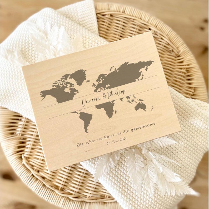 May include: A wooden world map with a gray silhouette of the continents. The names "Vanessa & Philipp" are written above a line, and below it, the text "Die sch&ouml;nste Reise ist die gemeinsame 26. Juli 2026" is displayed in a script font.