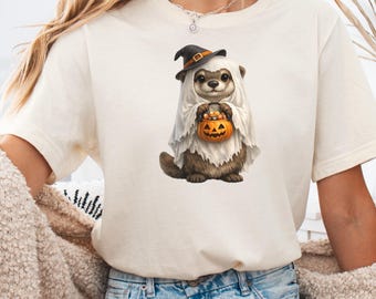 Può includere: Una t-shirt color crema con la grafica di una lontra marrone che indossa un costume da fantasma bianco e un cappello da strega nero. La lontra tiene tra le zampe anteriori un piccolo secchiello a forma di zucca arancione pieno di caramelle.