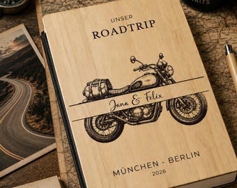 Può includere: Un taccuino rettangolare con copertina in legno chiaro caratterizzato da un'illustrazione incisa al laser di una motocicletta. Il testo riporta "UNSER ROADTRIP", "Jana & Felix", "MÜNCHEN - BERLIN" e "2026". Il taccuino ha il dorso nero e pagine bianche.