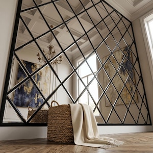 May include: Large, geometric wall mirror with a black metal frame and diamond-shaped panes. A woven basket with a cream-colored throw blanket sits in front of the mirror. The mirror reflects a room with art and a chandelier.