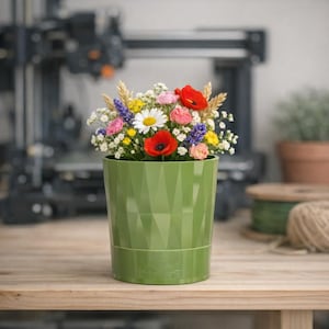 Peut inclure: Un pot de fleurs cylindrique en plastique vert olive avec une texture de surface géométrique à facettes. Le pot est rempli d'un bouquet varié de fleurs, comprenant des coquelicots rouges, des marguerites blanches, des roses roses et du blé.