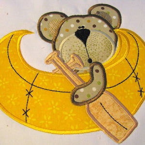 May include: A brown and tan embroidered bear with black eyes is sitting in a yellow crescent moon with a brown and tan embroidered paddle. The moon has a yellow floral pattern and is stitched with black thread.