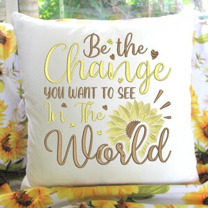 May include: White pillow with embroidered text "Be the Change You Want to See in the World" with a sunflower design. The pillow is on a floral fabric with a sunflower pattern.
