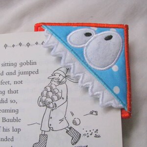 Könnte beinhalten: Ein blaues und weißes Stoff-Lesezeichen mit einem weißen gewellten Rand und einem roten Rand. Das Lesezeichen zeigt eine weiße Applikation eines Schneemanns mit zwei schwarzen Augen und einer roten Nase. Das Lesezeichen liegt auf einer Seite eines Buches mit Text über einen Kobold.