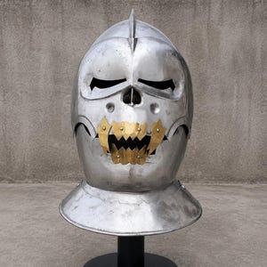 May include: A silver-colored metal helmet featuring a skull-like design with a central ridge, horizontal eye slits, and a prominent gold-colored metal jaw piece with jagged teeth attached to the front, mounted on a black circular stand.