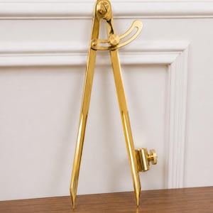 May include: A polished gold-toned metal drafting compass featuring two straight legs, a central pivot screw, an adjustable curved locking arm with a knurled nut, and a cylindrical attachment holder with a side-mounted tightening screw on one leg.