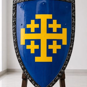 Könnte beinhalten: Ein wappenförmiger Schild mit glänzend blauer Oberfläche und einem großen gelben Jerusalemkreuz in der Mitte. Der Schild besitzt einen dunklen, verzierten Metallrand mit floralen Mustern, der mit kleinen, gleichmäßigen silbernen Nieten befestigt ist.