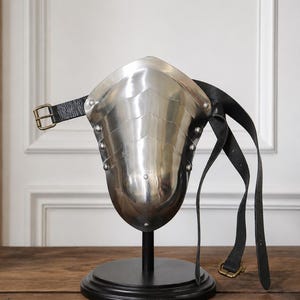 May include: A polished steel knee cop or poleyn armor piece featuring a multi-layered articulated design with visible rivets. The metal armor is secured by black leather straps with brass-toned buckles, mounted on a black circular display stand.