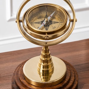 May include: A brass gimbal compass mounted on a tiered brass pedestal and a circular dark wood base. The compass features a glass cover, a central needle, and a dial marked with cardinal directions and degree increments in black lettering.