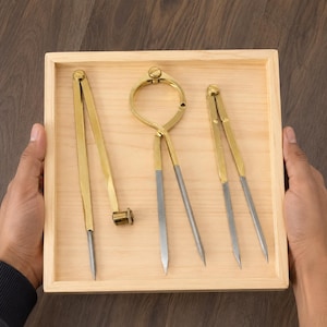 Könnte beinhalten: Ein Set aus drei Messzirkeln und Greifzirkeln aus Messing und Stahl, angeordnet in einem hellen Holztablett. Die Werkzeuge verfügen über goldfarbene Metallarme mit Stahlspitzen, darunter ein Zirkel mit einer Schraubverstellung.
