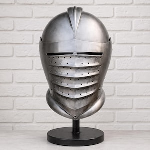 May include: A polished steel medieval-style knight helmet featuring a rounded dome with a central ridge, a horizontal eye slit, and a hinged visor with multiple rows of small ventilation holes, mounted on a black circular pedestal stand.