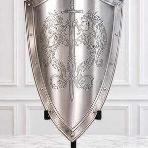 Könnte beinhalten: Ein silberfarbener Metallschild mit einer zentralen Gravur eines vertikalen Schwerts, das von zwei mythischen, drachenähnlichen Kreaturen flankiert wird. Der Schild hat einen verstärkten Rand mit sichtbaren Nieten und steht auf einem schwarzen Ständer.