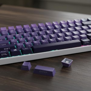 May include: A mechanical keyboard with purple keycaps and a white frame. The keycaps are a gradient of purple, with white lettering. The keyboard has a standard layout, with a few keycaps removed, showing the switches.
