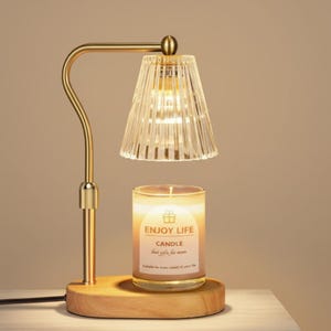 Könnte beinhalten: Ein Kerzenwärmer mit einem goldfarbenen Metallrahmen und einem transparenten, gerippten Glasschirm. Der Sockel ist aus Holz, und eine brennende Kerze befindet sich darunter. Die Kerze trägt die Aufschrift "Enjoy Life Candle".