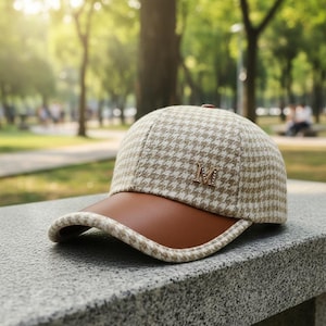 Puede incluir: Gorra de béisbol beige y marrón con estampado de pata de gallo y visera de cuero marrón. La gorra presenta la letra "M" dorada en la parte delantera. El sombrero está sobre una superficie de piedra.