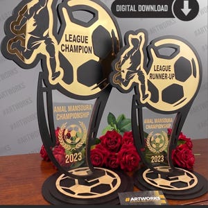 Puede incluir: Dos trofeos en negro y dorado con la silueta de un jugador de fútbol y un balón. El trofeo más grande dice LEAGUE CHAMPION y el más pequeño LEAGUE RUNNER-UP. Ambos muestran AMAL MANSOURA CHAMPIONSHIP 2023 con un icono de balón de fútbol.