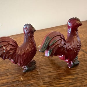 May include: Two matching cabinet knobs shaped like roosters, finished in a deep maroon red with dark, weathered accents on the tail feathers and crests. Each rooster figure features a textured surface and a metal mounting screw on the back.