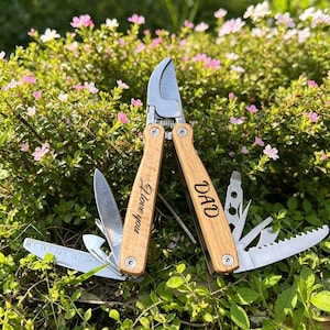 May include: A multi-tool garden pruner with wooden handles. One handle reads "I love you," the other "DAD." The tool includes a blade, saw, and ruler. The pruner is open, displaying its various functions.
