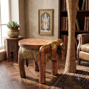 May include: A round wooden side table shaped like an elephant with a brown finish and gold-coloured decorative accents on the head and sides. The tabletop features a circular floral carving in the centre, and the base is supported by four legs.