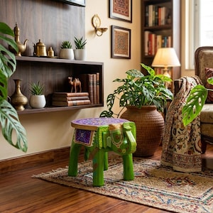 May include: A small, green, hand-painted wooden stool shaped like an elephant with a circular top featuring a purple and gold floral pattern. The stool has four sturdy legs and decorative carvings on the elephant's head, ears, and body.