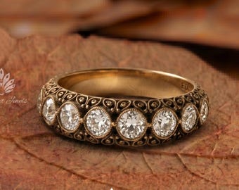 Puede incluir: Un anillo de banda dorada con una serie de piedras redondas, claras y facetadas dispuestas en fila. La superficie metálica muestra intrincados patrones de filigrana oscura que rodean cada piedra, creando un aspecto texturizado y detallado.