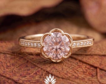 May include: A gold ring featuring a large, pale pink, flower-shaped gemstone. The gold band has small, clear stones set along the sides. The ring rests on a brown, textured surface. The ring is from Aerokoft Jewels.